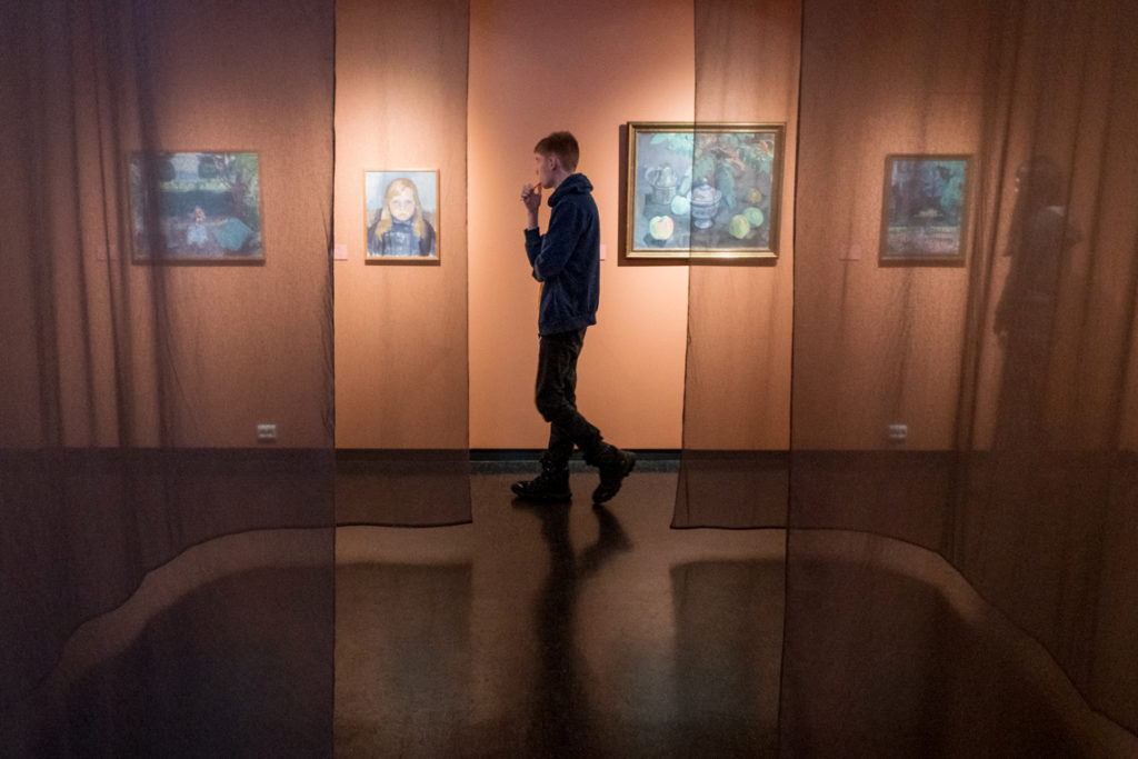 A room with paintings on the walls. A person walks behind a sheer drapery.